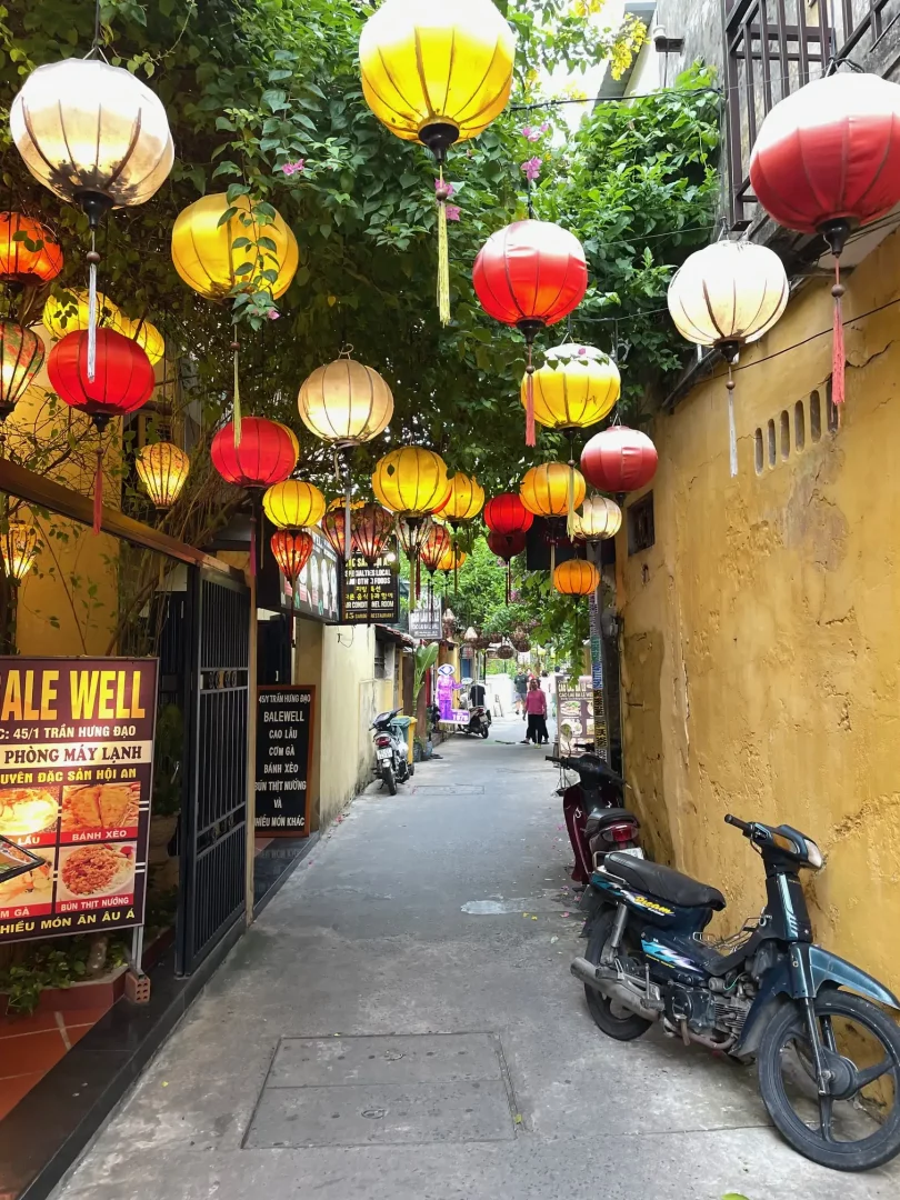 eine seitengasse bei tagelischt mit gelben hauwänden rechts und licks. über der gasse wachsen sträucher an denen bunte und farbenprächtige lampions hängen