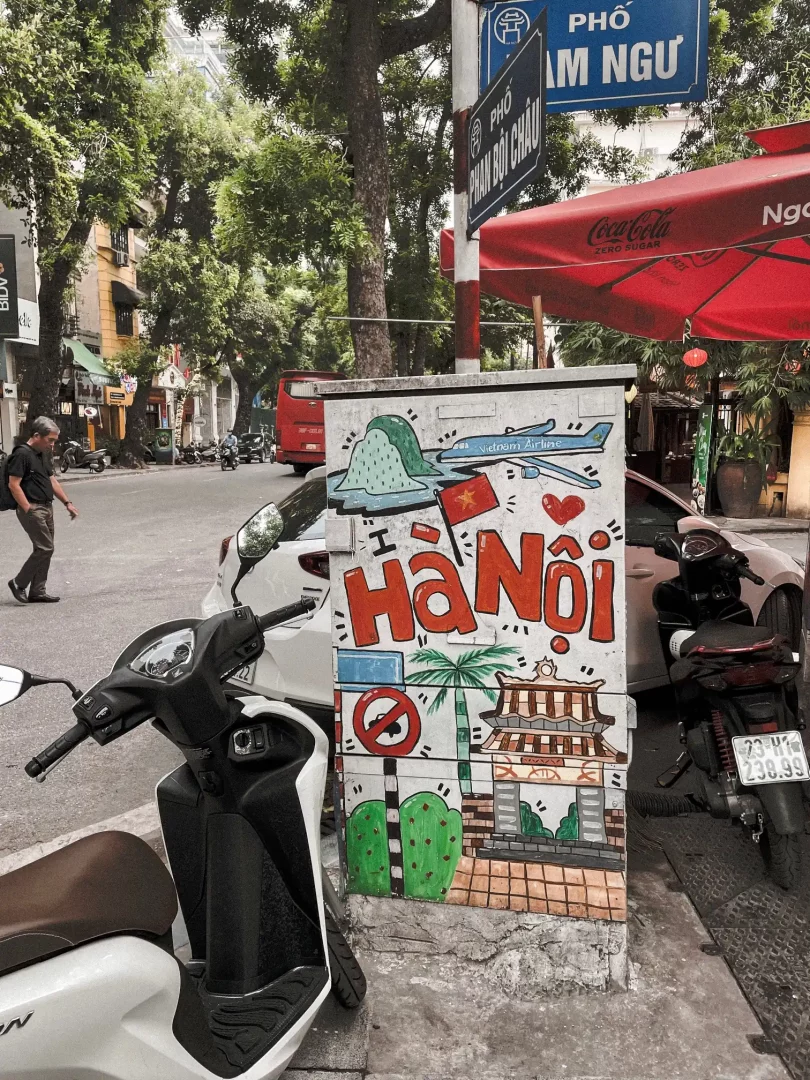 ein stromkasten bemalt mit der aufschrift hanoi in der farbe rot.davor stehend ein weißer motorroller